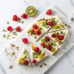 Sliced frozen yogurt bark with raspberries, orange slices, and pumpkin seeds on a marble board.